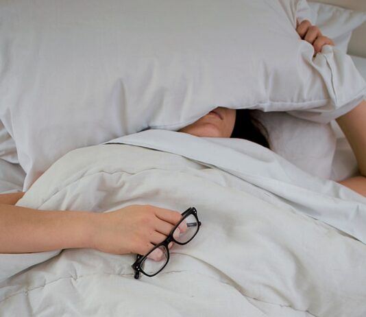 피로회복을 위한 에너지 충전 person lying on bed while covering face with pillow and holding eyeglasses